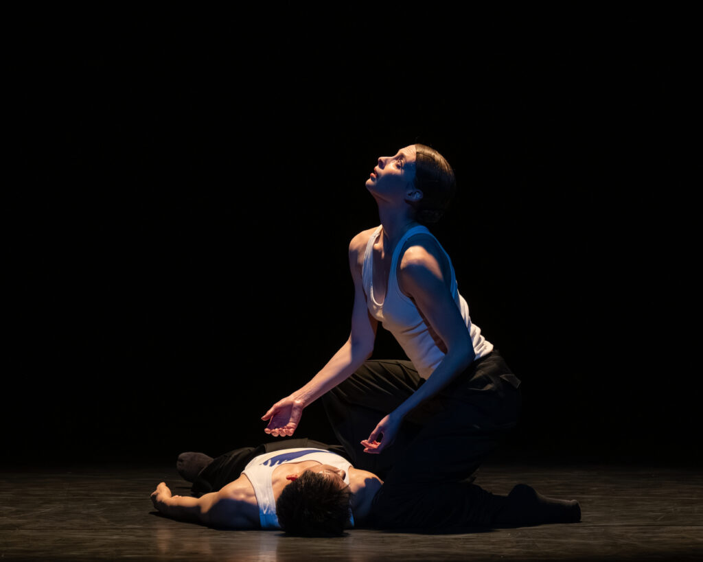 Emma Hawes kneelsm grieving over the body of Ken Saruhashi in the final scene of Cyrstal Pite's Body & Soul Part 1