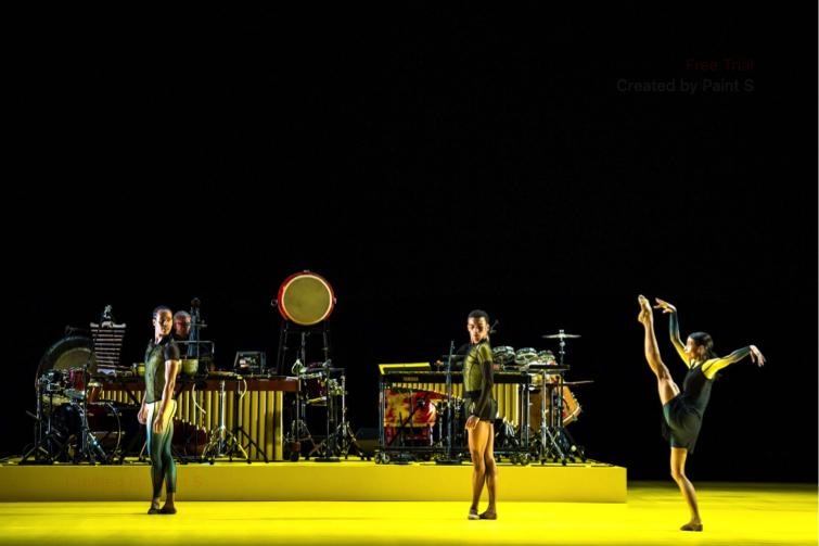 Two Royal Ballet male dancers watch as a female dancer raises one leg.  All stand on a yellow floor in front of a complicated percussion set, in Quantum Souls part of Wayne McGregor Alchemies