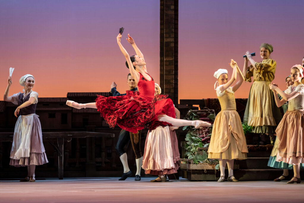 Kitri in a red flouncy dress flies across the stage in grand jeté, clutching a fan in Act I of Don Quixote