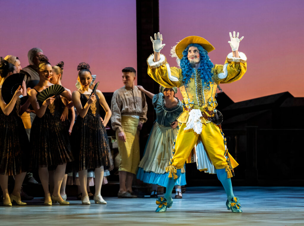 The foppish Gamache dressed in extravagant gold and blue, with white gloves, feathered hat and long blue wig