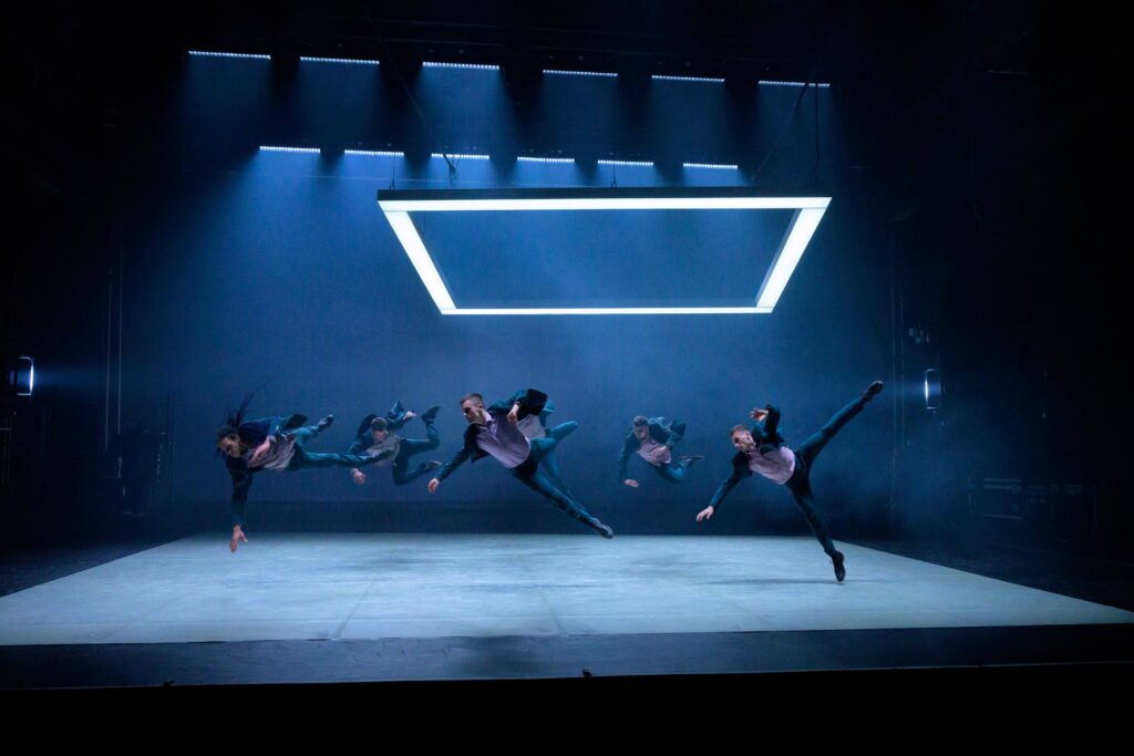 Five dancers jump on a blue-lit stage under a neon square