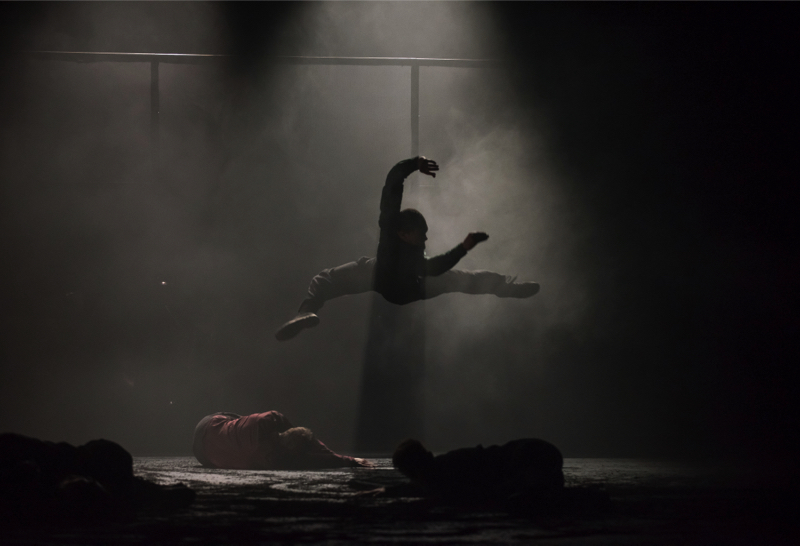A dancer leaps up against a dark a dusty background. Two others lie on the ground.