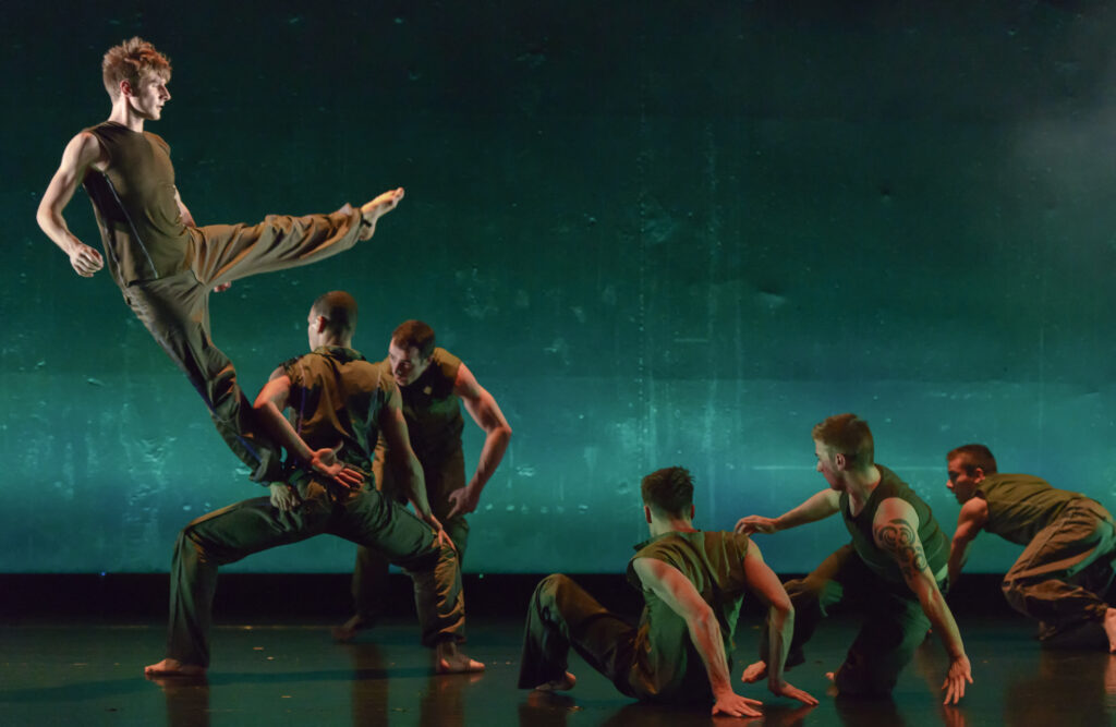 One dancer stands on another's thigh with one leg stretched out while four others prepare to rise off the ground