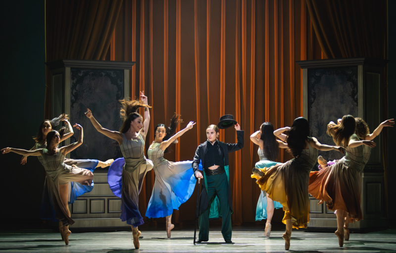 Gentleman Jack raises her top hat as she stands surounded by female dancers in flowing colourful skirts with loose hair