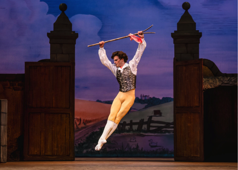 Colas, wearing yellow tights and a printed waistcoat over a white shirt, jumps with legs held together at an angle, while holding a forked stick with a pink ribbon tied on.
