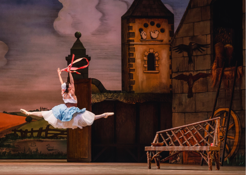 Lise jumps while holding a pink ribbon aloft in the yart of her house, where you can see a bench, with a tower in the background