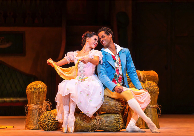 Lise, wearing a predominantly pink costume with embroidered skirt, holds a yellow neckerchief while looking lovingly at Colas, who sits next to her wearing yellow tights and light blue jacket and holding a pink neckerchief.