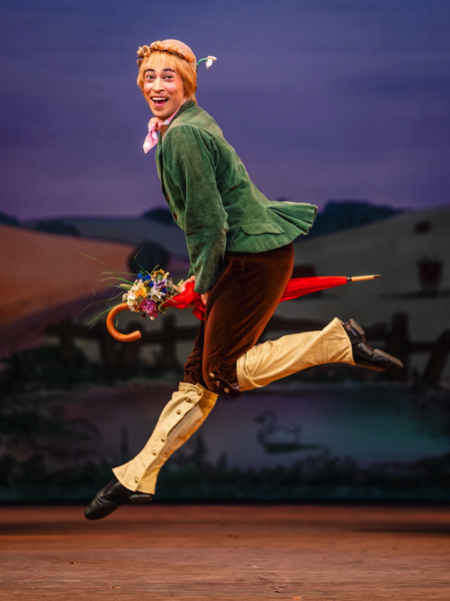 Alain, in brown velvet breeches , green jacket and yellow spats holds a small bouquet of flowers and rides his red umbrella grinning at the audience