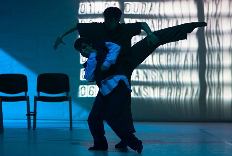 A couple dance in front of an airport departures board with the letters dissolving,  THe man facing us has one leg raised to the side, the woman embraces him.