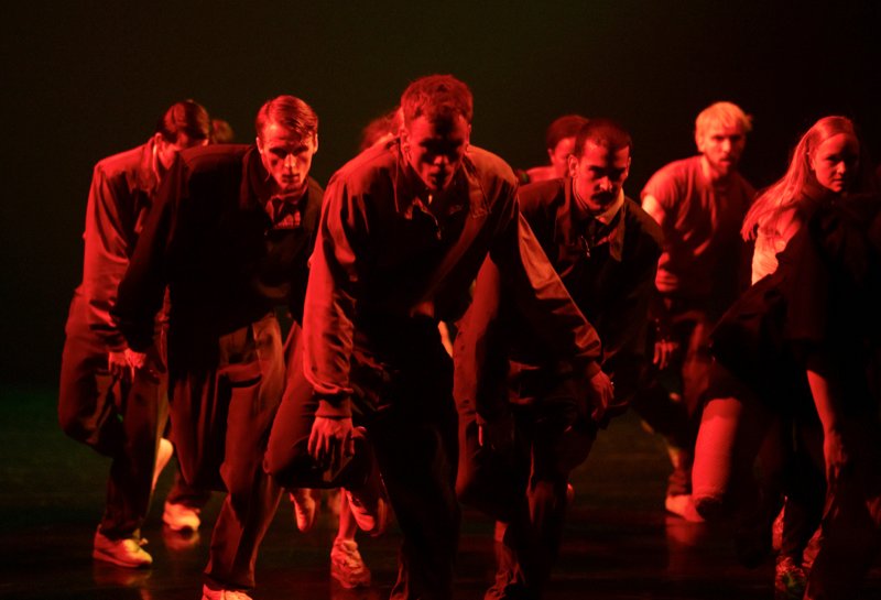 This is Rambert: a group of re lit dancers wearing street clothes lean forward with hunched shoulders.