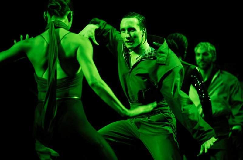The iamge is green lit and shows a male dancer facing a woman with bare arms and shoulders with her back to us