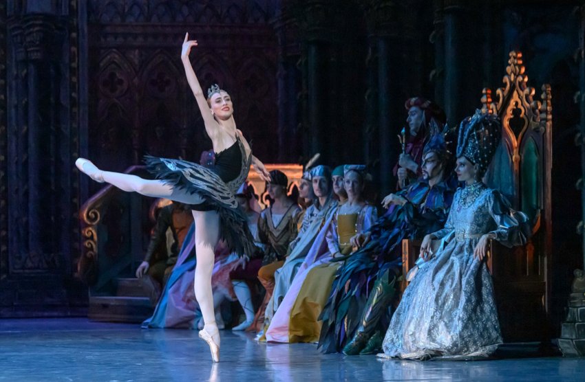 Swan Lake, Ball Scene: the queen, wearing an elaborate tall headdress, sits on a throne surrounded by her entourage, watching the Black Swan Odile, who stands on pointe on her left let, the right raised in attitude behind her, right arm raised above her head.