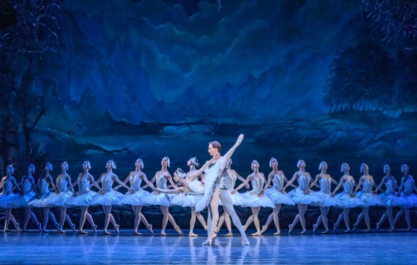 State Ballet of Georgia Swan Lake Act I Scene II by the lakeside. The prince supports the Swan princess as she stands on pointe with one leg lifted in high arabesque. They stand in front of a line of identically poised swan women