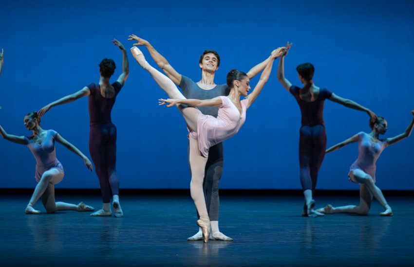 Three couples of Royal ballet School students. The two women at the back kneel looking away from the centre, white the men stand with their backs to us, one arm raised. The central couple pose with the pink dressed woman in high arabesque on pointe, the grey dressed man standing behind and lightly holding her hand