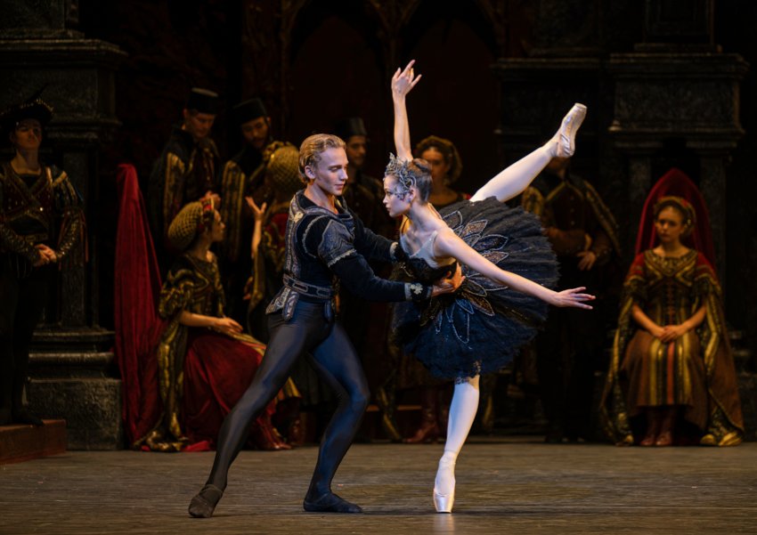 Royal Ballet Scjool students in Black Swan pas de deux. Both are dressed in black: the man supports the woman as the leans forward with her right leg in high attitude, arms to the sides like wings.
