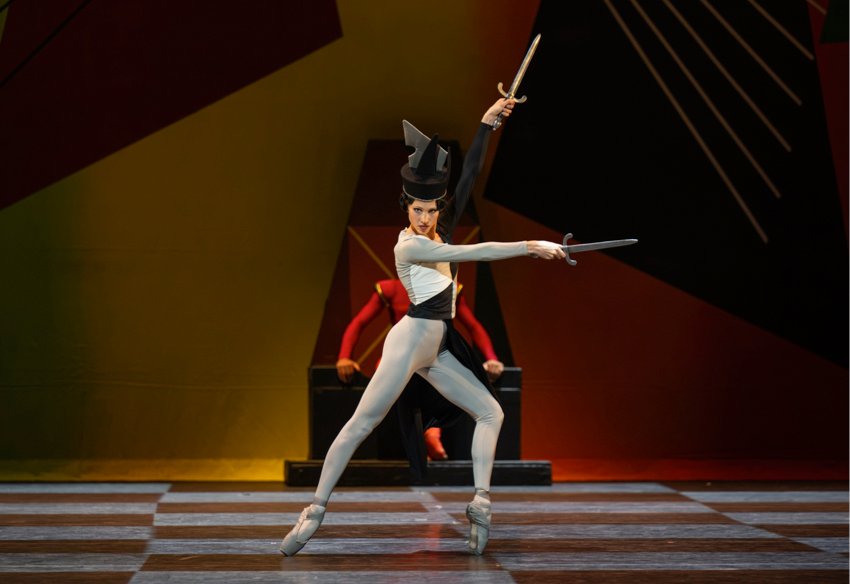 The Royal Ballet School student dances the Black Queen dressed in grey bodysuit with a geometric black empiecement and a tall black head dress, She stands on pointe, one leg bent, the other stretched behind and holds two swords