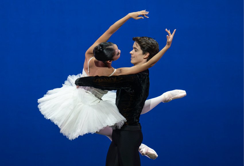 A man dressed in black lifts a woman in a white tutu, who has her back to us. Her legs are stretched forwards, arms raised as it to embrace him, and they look each other in the eyes smiling.