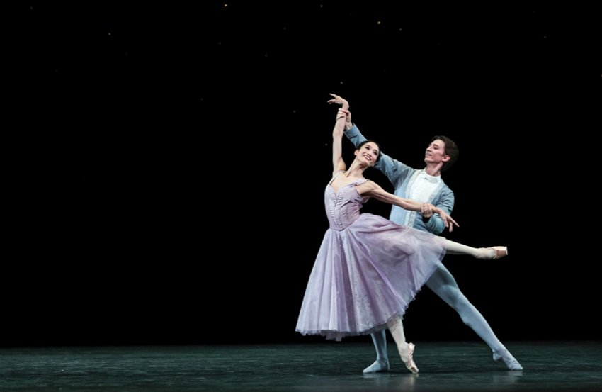 Marianela - Timeless. Fumi Kaneko wears a light lilac long skirt and stands on pointe on her right leg with the left raised behind in arabesque, left arm parallel, right arm raised. Muntagirov in soft blue, holds both her wrists softly.
