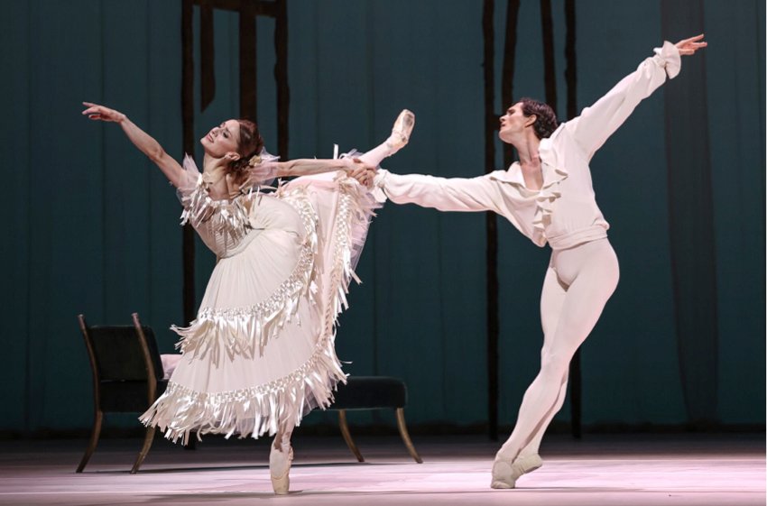 Núñez wearing full skirt with satin fringes stands on pointe on her left leg, right lifted behind in arabesque, left arm extended forward. Bracewell holds her right arm as if to pull her towards him. Both are dressed in white.