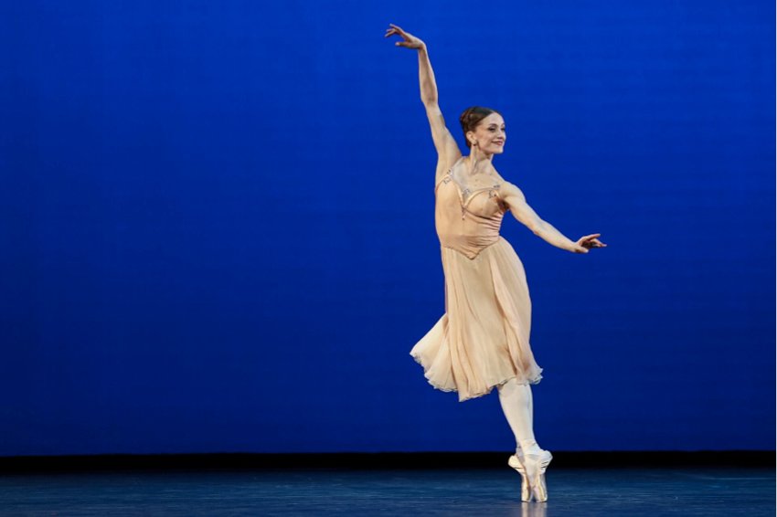Marianela - Timeless. Against a deep blue backdrop, Núñez stands on pointe on the right of the image, right arm raised, left extended to the side. She is weating a floaty gold dress and is smiling