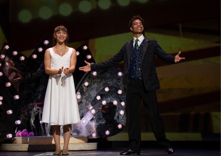 Myths and Modern Masters: Nutcracker in Havana Suite: young Clara in a sleeveless white dress claps while Carlos Acosta in a three piece suite, stands to her left smiling with arms open