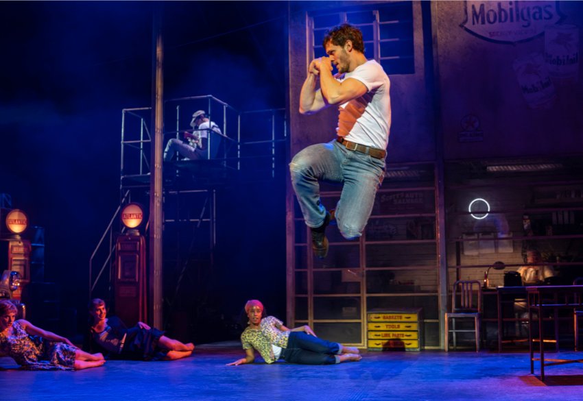 The Car Man: Luca, wearing jeans and a white T-shirt, jumps up with legs bent and hands clenched in front of his face, watched by three women lying down