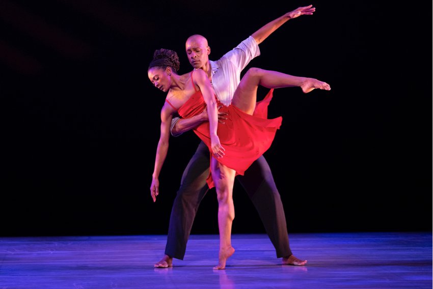 Alvin Ailey American Dance Theater A woman in a red dress stands on demi-pointe on her right leg, with the left raised in attitude to the side.  She is supported by a man in black trousera and a white shirt