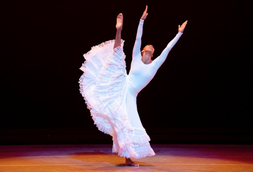 Alvin Ailey American Dance Theater: a woman in a long, ample, flouncy skirt raises her right leg up above her head, arms and head raised to heaven