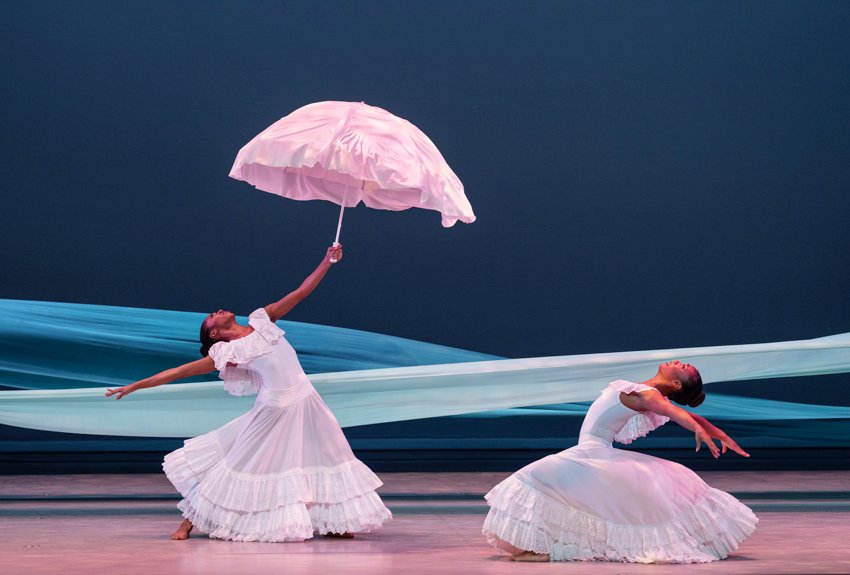 Alvin Ailey American Dance Theatre, Revelations. Against a dark blue background, two blue strips of cloth simulate a flowing river.  Two women in ample, flouncy white dresses stand, one holding a white parasol, both in deep backbends.