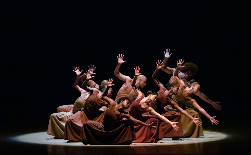 Alvin Ailey American Dance Theatre, Revelations. A cluster of dancers clad in deep earth tones, stand in deep knee bend leaning to our right, with the left arm stretch sideways, the right raise up, fingers splayed