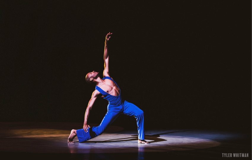 Alvin Ailey American Dance Theater, A Song For You.  A man in a deep blue body suit with a plunging neck lline stands on a bent knee, his right leg behind, left arm and head lifted high.