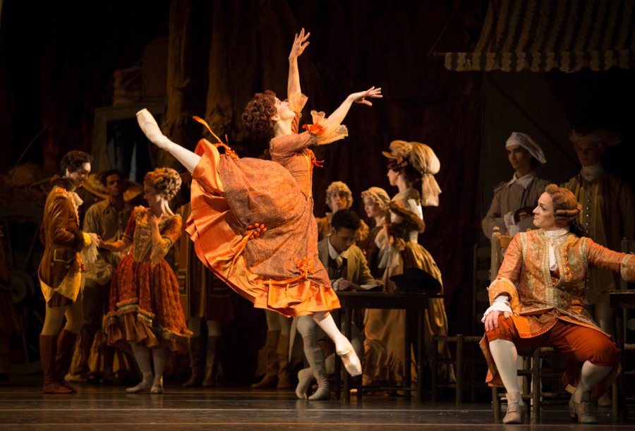 Mayara Magri as Lescaut's Mistress in Manon, dressed in a rich orange costume, leaps in arabesque observed by her patron Monsieur GM, who's sitting to our right