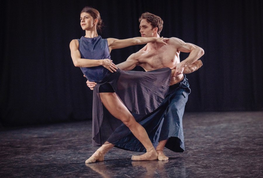 Mayara Magri and Matthew Ball dance a contemporary piece.  They wear indigo costumes. On bent knees he holds her in an off balance arabesque